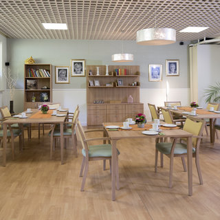 Ein heller Speisesaal mit mehreren Tischen und Stühlen. Jeder Tisch ist gedeckt mit Tellern und Besteck. An den Wänden hängen Bilder, und im Hintergrund steht ein Regal mit Büchern und Pflanzen. Der Raum hat eine angenehme, einladende Atmosphäre.