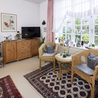 Ein gemütliches Wohnzimmer mit zwei Korbstühlen und einem kleinen Tisch, auf dem Pflanzen stehen. An der Wand hängt ein Bild, und in der Ecke steht ein Fernseher. Große Fenster lassen viel Licht herein und bieten Ausblick auf den Außenbereich. Der Boden ist mit einem Teppich bedeckt.