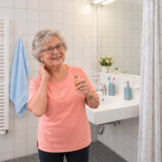 Eine ältere Frau mit grauen Haaren und Brille lächelt, während sie in einem Badezimmer steht. Sie hält ein Parfümfläschchen in der Hand. Im Hintergrund sind ein Waschbecken, eine Dusche und Badezimmeraccessoires sichtbar.