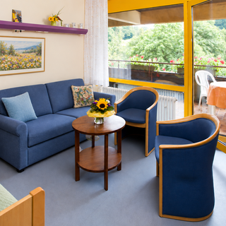 Ein helles, einladendes Wohnzimmer mit blauen Möbeln. Ein Sofa mit Kissen und zwei Stühlen um einen runden Tisch. Große Fenster bieten Blick auf einen Balkon mit Blumen und einem Tisch. Künstliche Pflanzen und ein Gemälde schmücken die Wände.