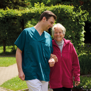 Ein junger Mann in einem tealfarbenen medizinischen Outfit und eine ältere Frau in einem roten Fleece-Jacke gehen lächelnd Hand in Hand in einem Garten. Im Hintergrund sind grüne Sträucher und ein sonniger Himmel zu sehen.