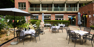 Ein älterer Mensch mit einem Rollator steht in einem Innenhof, umgeben von Tischen mit Stühlen und Sonnenschirmen. Die Tische sind mit weißen Tischdecken gedeckt. Im Hintergrund ist ein mehrstöckiges Gebäude zu sehen, das eine einladende Atmosphäre schafft.