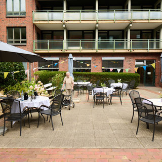 Ein älterer Mensch mit einem Rollator steht in einem Innenhof, umgeben von Tischen mit Stühlen und Sonnenschirmen. Die Tische sind mit weißen Tischdecken gedeckt. Im Hintergrund ist ein mehrstöckiges Gebäude zu sehen, das eine einladende Atmosphäre schafft.