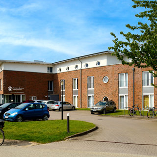 Ein modernes Seniorenheim aus rotem Ziegelstein. Vor dem Gebäude stehen mehrere Autos und Fahrräder. Eine Person auf einem Fahrrad ist im Vordergrund sichtbar, während im Hintergrund einige Bäume zu sehen sind. Der Himmel ist klar und blau.