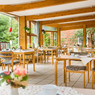 Ein hell erleuchteter Speisesaal mit großen Fenstern, die auf einen grünen Außenbereich blicken. Holztische sind ordentlich deckiert, einige mit Blumendekoration. Die Atmosphäre wirkt einladend und freundlich, ideal für Mahlzeiten und gesellige Zusammenkünfte.