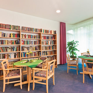 Der Raum zeigt eine gemütliche Bibliothek mit einem Bücherregal vollgepackt mit Bücher. Es gibt mehrere Tische mit Stühlen, die einladend wirken. Ein grüner Tisch ist dekoriert mit einer kleinen Blumenarrangierung. Ein Fenster mit Vorhängen lässt natürliches Licht herein, und eine Pflanze steht in der Ecke.