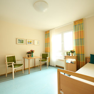 Das Bild zeigt ein helles Zimmer mit einem Einzelbett, einem Tisch und zwei Stühlen. An der Wand hängen Bilder, und auf dem Tisch steht eine kleine Pflanze. Vor einem Fenster hängen gestreifte Vorhänge. Der Boden ist blau.