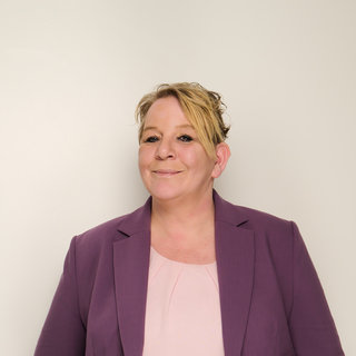 Eine lächelnde Frau steht mit den Händen in den Taschen ihrer Jeans. Sie trägt ein pinkes Oberteil und einen lila Blazer. Der Hintergrund ist hell und neutral gehalten.