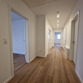 Ein heller Flur mit Laminateboden und weißen Wänden. Von beiden Seiten sind Türen sichtbar, die in verschiedene Räume führen. An der Decke sind helle LED-Leuchten angebracht, die den Flur gut ausleuchten. Der Raum vermittelt einen modernen und offenen Eindruck.