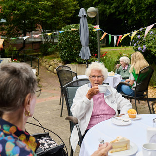 Eine Gruppe älterer Frauen sitzt draußen in einem Gartenlokal. Einige genießen Kaffee und Kuchen an einem Tisch, während Buntgirlanden im Hintergrund hängen. Die Atmosphäre ist entspannt und gesellig, mit Pflanzen und Pflanzenkübel um sie herum.