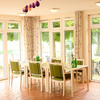 Ein hell erleuchteter Speiseraum mit mehreren Tischen und Stühlen. Große Fenster bieten einen Blick auf einen grünen Außenbereich. Im Hintergrund ist eine Person im Rollstuhl sichtbar. Die Wände sind mit Vorhängen dekoriert, und die Tische sind für eine Mahlzeit gedeckt.