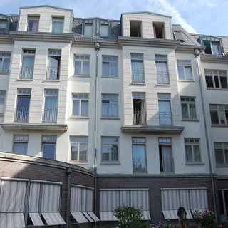 Ein mehrstöckiges, helles Wohngebäude mit weißen Wänden und mehreren Fenstern. Im unteren Bereich sind Fenster mit Markisen und ein kleiner Garten mit Blumen sichtbar. Der Himmel ist blau mit einigen Wolken.