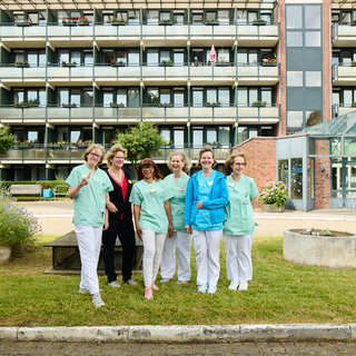 Sechs Pflegekräfte stehen gemeinsam in einem Außenbereich vor einem Wohngebäude. Sie tragen unterschiedliche Arbeitskleidung in hellen Farben und lächeln in die Kamera. Im Hintergrund sind Balkone und Pflanzen sichtbar.