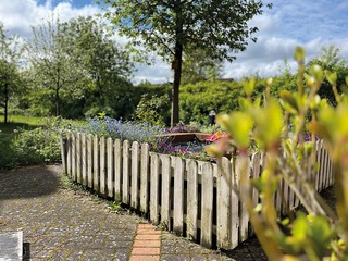 Der Boden zeigt einen gemauerten Bereich mit einem weißen Lattenzaun, der ein Blumenbeet umgibt. Bunte Blumen wachsen darin, während im Hintergrund ein Baum und grüne Wiesen sichtbar sind. Der Himmel zeigt einige Wolken und blauen Himmel.