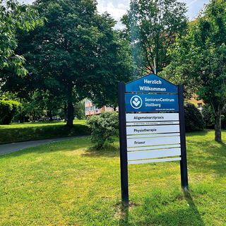 Ein Begrüßungsschild des SeniorenCentrum Stolberg in einer grünen Umgebung. Das Schild enthält Informationen über eine Allgemeinarztpraxis, Physiotherapie und einen Friseur. Im Hintergrund sind Bäume und ein Gehweg sichtbar.