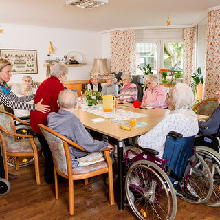 Eine Gruppe älterer Menschen sitzt um einen Tisch in einem Gemeinschaftsraum. Einige sind im Rollstuhl, während andere auf Stühlen sitzen. Eine Betreuerin spricht mit einer Teilnehmerin. Die Atmosphäre wirkt freundlich und gesellig. Es gibt Getränke und Snacks auf dem Tisch.