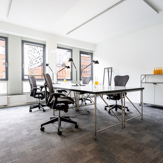 Büro mit mehreren Stühlen und einem großen Tisch in der Mitte. Am Tisch stehen Lampen und einige Flaschen mit Getränken. Große Fenster lassen natürliches Licht herein und bieten Blick auf Stadtgebäude. Der Boden ist mit Teppich ausgelegt.