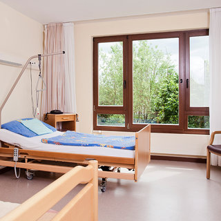 Ein Patientenraum mit einem Krankenhausbett, das mit einer blauen Decke und Kissen ausgestattet ist. Neben dem Bett befindet sich ein Nachttisch. Große Fenster bieten Blick auf einen kleinen Garten mit Bäumen und Grün. Der Raum ist hell und hat eine freundliche Atmosphäre.