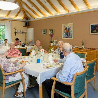 Eine Gruppe älterer Menschen sitzt an einem Tisch in einem hellen Raum. Sie genießen eine Mahlzeit und unterhalten sich. Um den Tisch herum sind Speisen und Getränke präsentiert. Im Hintergrund sind Fenster, die Tageslicht und eine grüne Aussicht hineinlassen.
