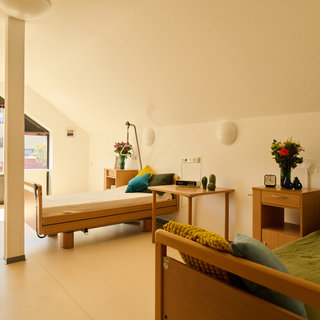 Ein helles Zimmer mit zwei Krank betten, einem Fenster mit Ausblick und einer modernen Einrichtung. Auf den Betten liegen Kopfkissen und Decken. Es gibt einen Tisch mit Pflanzen und eine Stehlampe. Der Raum vermittelt eine freundliche, einladende Atmosphäre.