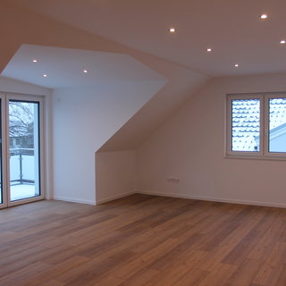 Ein helles, modernes Zimmer mit Holzfußboden und weißen Wänden. Große Fenster lassen viel Tageslicht hereinfällen. Eine Wand hat eine schräge Form, und auf dem Balkonen ist frischer Schnee zu sehen. Die Decke ist mit mehreren integrierten Lichtern beleuchtet.