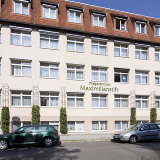 Das Bild zeigt die Fassade eines Pflegezentrums mit dem Namen "Maximilianstift". Es handelt sich um ein mehrstöckiges Gebäude mit mehreren Fenstern und einer begrünten Umgebung. Parkplätze sind im Vordergrund sichtbar.