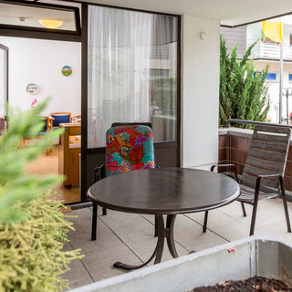 Ein Gartenbalkon mit einem runden Tisch und zwei Stühlen. Einer der Stühle hat eine bunte Sitzauflage. Im Hintergrund ist ein Raum mit Möbeln sichtbar. Üppige Pflanzen umgeben den Bereich, und es gibt einen Blick auf eine Straße mit einem roten Auto.