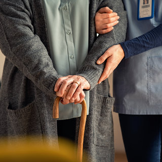 Eine ältere Person steht mit einem Gehstock, den sie festhält. Eine Pflegerin unterstützt sie, indem sie ihren Arm sanft umlegt. Die Szene vermittelt Fürsorge und Unterstützung in einem häuslichen Umfeld.