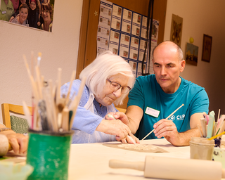 Eine ältere Frau mit Brille und einem Mann arbeiten gemeinsam an einer Tontafel. Der Mann leitet die Frau beim Arbeiten, während kreative Materialien wie Pinsel und Farbbehälter auf dem Tisch liegen. Im Hintergrund sind Bilder an der Wand sichtbar.