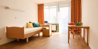 Das Bild zeigt ein helles, schlicht eingerichtetes Zimmer mit einem Einzelbett, einem Tisch mit rotem Stuhl und einem kleinen Beistelltisch. Vor einem großen Fenster hängen orangene Vorhänge, und es gibt ein paar Dekorationsgegenstände auf dem Tisch. Der Boden ist aus Holz.