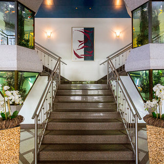 Eine elegante Treppe führt in einem hell erleuchteten Foyer nach oben. An beiden Seiten stehen Blumengebinde mit Orchideen, und geschwungene Geländer rahmen die Treppe ein. Über der Treppe hängt ein modernes Kunstwerk.