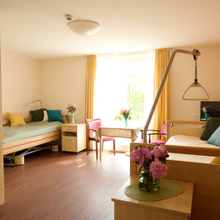 Ein helles Zimmer mit Holzdekor, zwei Betten, einem Tisch und Stühlen. Auf einem Tisch stehen frische Blumen. Ein Fenster mit Vorhängen sorgt für Tageslicht, das den Raum freundlich wirken lässt. Die Atmosphäre ist einladend und gemütlich.