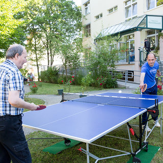 Zwei Männer spielen Tischtennis im Freien, umgeben von Bäumen und Blumen. Die eine Person trägt ein kariertes Hemd, während die andere ein blaues T-Shirt anhat. Im Hintergrund ist ein Gebäude mit einer Eingangsveranda zu sehen.