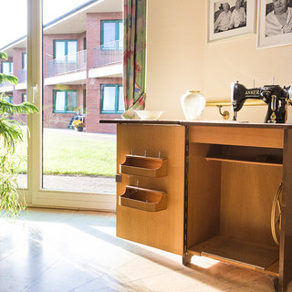 Ein Holztisch steht im Raum und enthält eine Nähmaschine. Daneben sind zwei Schubladen sichtbar. Hinter dem Tisch gibt es große Fenster mit Blick auf einen Rasen und Gebäude. Eine Pflanze in einem Korb ist im Vordergrund zu sehen.