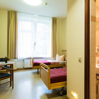 Ein einfaches Zimmer mit zwei Krankenbetten, einem Stuhl und einem kleinen Tisch. Es gibt ein Fenster mit Vorhängen, das Tageslicht hereinlässt. Im Hintergrund ist ein Badezimmer mit einer Badewanne sichtbar. Die Wände sind hell und der Boden ist neutral gehalten.