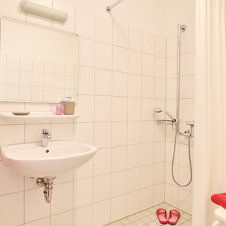 Das Bild zeigt ein Badezimmer mit weißer Fliese. Es gibt eine Dusche mit Duschvorhang, ein Waschbecken mit Spiegel darüber und eine kleine Ablage. Rote Handtücher hängen neben dem Waschbecken, und es stehen rosa Hausschuhe am Boden.