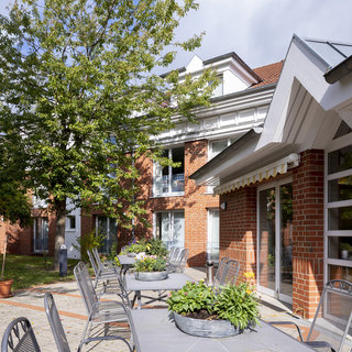 Ein Außenbereich mit mehreren Tischen und Stühlen, umgeben von Bäumen und Pflanzen. Im Hintergrund sind Gebäude mit großen Fenstern sichtbar. Die Szene strahlt eine ruhige, einladende Atmosphäre aus, perfekt für Entspannung im Freien.
