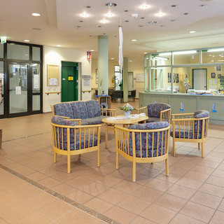 Eine helle Empfangshalle mit einem Wartebereich. In der Mitte stehen mehrere Korbstühle um einen Tisch, dekoriert mit einer kleinen Blumenvase. An einer Wand befindet sich ein Empfangstresen mit transparenten Fenstern und Informationsmaterialien. Der Boden ist gefliest.