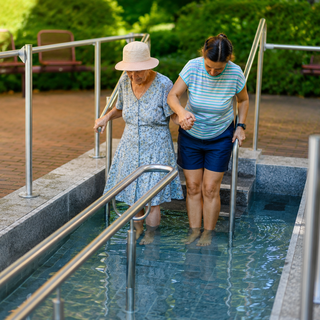 Eine ältere Frau in einem blauen Blumenkleid mit Sonnenhut geht langsam zusammen mit einer jüngeren Frau in einem gestreiften T-Shirt und Shorts in ein warmes Wasserbecken. Beide halten sich aneinander fest und scheinen die angenehmere Temperatur zu genießen.