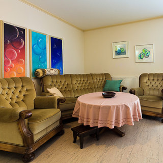 In einem hellen Wohnzimmer stehen zwei grüne Sofas und ein runder Tisch mit einer pinkfarbenen Tischdecke. Auf dem Tisch befindet sich eine Schale. An der Wand hängen farbenfrohe abstrakte Bilder und zwei weitere Rahmen mit Kunstwerken.