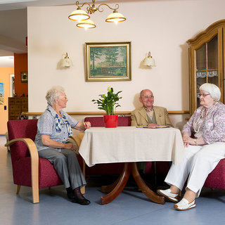 Drei ältere Menschen sitzen an einem Tisch in einem Aufenthaltsraum. Eine Frau mit grauem Haar trägt eine Brille, ein Mann in einem hellen Anzug und eine weitere Frau mit blonden Haaren unterhalten sich. Auf dem Tisch steht ein Topf mit einer Pflanze.