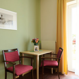 Ein ruhiger Raum mit grün gestrichenen Wänden. In der Ecke steht ein runder Tisch mit zwei Stühlen, die rote Polster haben. Auf dem Tisch befindet sich eine Blumenvase und ein gerahmtes Foto. Ein Fenster mit Vorhängen lässt Licht herein.