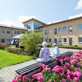 Zwei ältere Menschen sitzen auf einer Bank im Außenbereich eines Wohnheims. Um sie herum blühen farbenfrohe Blumen, und im Hintergrund sind die Fenster eines Gebäudes zu sehen. Der Himmel ist klar und die Atmosphäre wirkt ruhig und einladend.