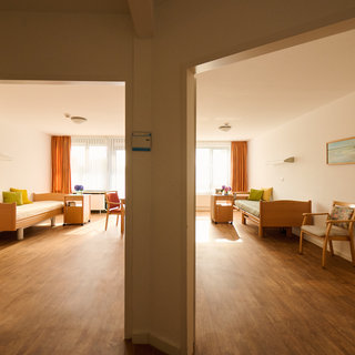 Die Abbildung zeigt einen hellen Flur mit zwei offenen Zimmern. Jedes Zimmer ist einfach eingerichtet mit einem Bett, kleinen Tischen, Stühlen und Fenstern, die Tageslicht hereinlassen. Die Wände sind in neutralen Farben gehalten, und es gibt eine warme Atmosphäre.