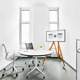 Ein helles, minimalistisches Büro mit weißen Wänden. In der Mitte steht ein runder Tisch mit einem Stuhl. Auf dem Tisch liegt ein Notizblock. An einer Wand steht ein Flipchart mit Diagrammen, daneben stehen Getränke in einer Kühlbox.