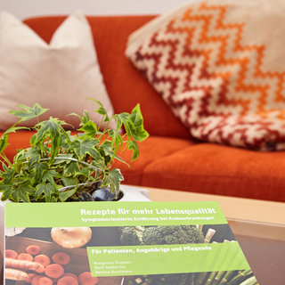 Ein Tisch mit einem Kochbuch mit dem Titel „Rezepte für mehr Lebensqualität“ und einer kleinen Pflanze. Der Hintergrund zeigt ein oranges Sofa mit einem gemusterten Kissen. Das Buch richtet sich an Patienten, Angehörige und Pflegende.