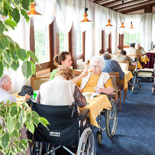 Eine helle, freundliche Speiseräume mit Erwachsenen in Rollstühlen, die an Tischen sitzen. Eine Betreuerin füttert eine ältere Dame, während andere Gäste miteinander sprechen. Große Fenster lassen Tageslicht herein und Pflanzen dekorieren den Raum.