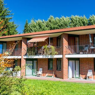 Ein modernes Wohngebäude mit mehreren Etagen, das aus roten Ziegeln besteht. Jede Etage hat Balkone, einige mit Pflanzen dekoriert. Im Vordergrund sind Grünflächen zu sehen, die das Gebäude umgeben. Der Himmel ist klar und blau.