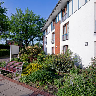 Ein Mehrfamilienhaus mit modernen Fenstern und einem begrünten Vorgarten. Der Garten ist mit verschiedenen Pflanzen gestaltet, darunter Sträucher und Blumen. Eine Holzbank steht auf dem gepflasterten Weg vor dem Haus. Im Hintergrund sind Bäume sichtbar.