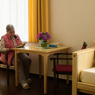 Eine ältere Frau sitzt an einem Tisch in einem hellen Raum und liest ein Buch. Auf dem Tisch steht eine Vase mit frischen Blumen. Die Wände sind in neutralen Tönen gehalten, und es gibt einen Stuhl und eine Liege im Raum. Sonnenlicht fällt durch das Fenster.